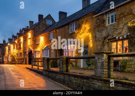 Luci di Natale di notte al di fuori delle case e il Crown hotel nella strada alta. Blockley, Cotswolds, Gloucestershire, Inghilterra Foto Stock