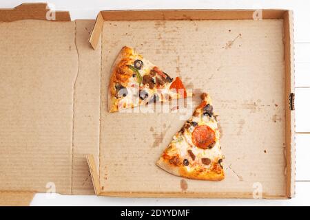 Closeup open box con due fette di pizza con pepperoni, carne tritata, funghi, pepe dolce e olive su un tavolo di legno bianco. Vista dall'alto. Foto Stock