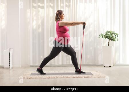 Foto di profilo di lunghezza completa di una donna incinta che si esercita con una fascia yoga in gomma resistente su un tappetino a casa Foto Stock