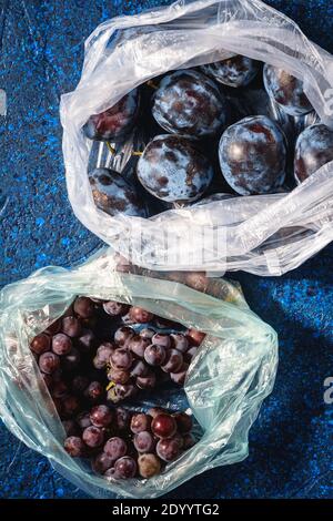 Frutti di prugne e acini d'uva freschi maturi in confezione di sacchetti di plastica su sfondo astratto blu, vista dall'alto Foto Stock