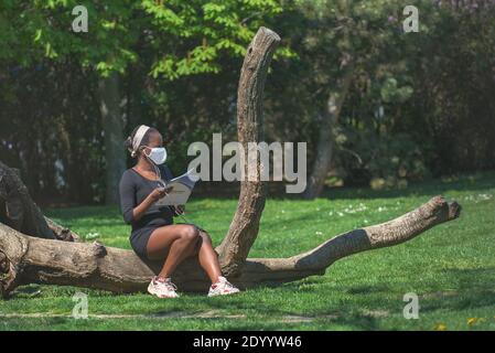 Donna afroamericana con una maschera di stoffa seduta su un tronco di albero sdraiato nel parco vicino alla coda per il test del virus corona. Foto Stock