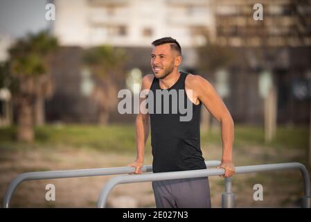 Calzare il giovane uomo facendo tricipite immerge su barre parallele all'aperto. Fare esercizi su barre parallele nel parco Foto Stock