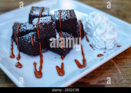 Una porzione di brownie con una fonte di gocciolamento di caramello dolce e panna bianca servita su un piatto bianco con zucchero in polvere. Foto Stock