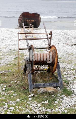 Una piccola vecchia barca da pesca in legno aperta collegata con filo ad un argano arrugginito, con il mare sullo sfondo sulla riva di ciottoli. Foto Stock