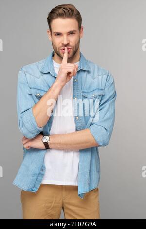 il giovane bell'uomo che indossa una camicia in jeans tiene il dito sulle labbra, chiede di non comunicare informazioni segrete o di mantenere il silenzio Foto Stock