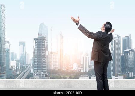 Vista posteriore di un uomo d'affari asiatico in piedi con la mano sollevata il tetto con i grattacieli sullo sfondo Foto Stock