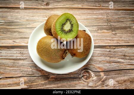 Frutta intera di kiwi matura e metà di kiwi in piatto bianco su fondo di legno. Profondità completa di campo. Concetto di cibo vegano Foto Stock