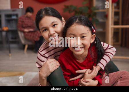 Ritratto medio ravvicinato di due giocose ragazze asiatiche che si divertono insieme in salotto sorridendo alla macchina fotografica mentre la loro madre lavorare su un computer portatile Foto Stock