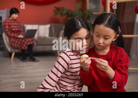 Due ragazze cinesi curiose che si siedono sul pavimento nel soggiorno che legge il messaggio di previsione del biscotto di fortuna, spazio di copia Foto Stock