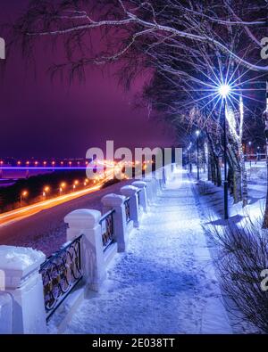 Parcheggio invernale di notte con lanterne, vista su strada con moto d'auto, marciapiede e alberi ricoperti di neve. Foto Stock
