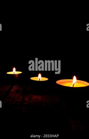 Tre candele di cera da tè illuminate su un tavolo di legno su sfondo nero. Foto Stock