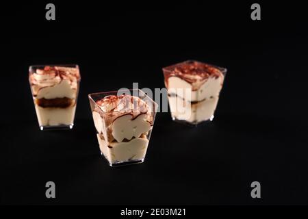 Torta tiramisù con tre strati di biscotto al cioccolato e sciroppo di caffè naturale con cognac e crema. Tre bicchieri a fila decorati con panna montata e cacao, isolati su sfondo nero. Foto Stock