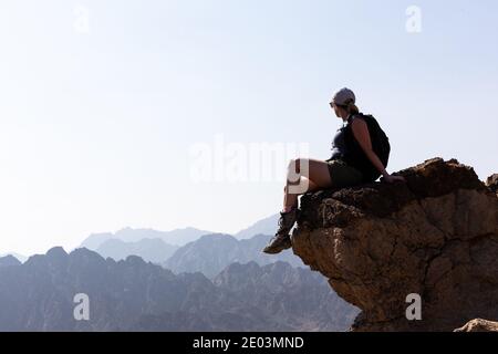 Giovane donna turistica seduto al bordo della roccia con abisso sotto, guardando le creste di montagna all'orizzonte, Hatta, Hajar Mountains Foto Stock