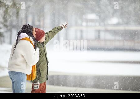 Giovane coppia in abiti caldi che guarda qualcosa in piedi all'aperto quando nevica Foto Stock
