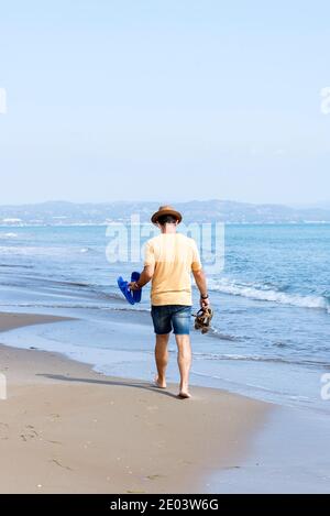 Vista posteriore dell'uomo con cappello, con le scarpe che si tengono a piedi nudi in riva al mare Foto Stock