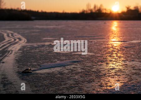 Arancione brillante tramonto su ghiacciata superficie del lago riflettente ghiacciato primo piano con alberi scuri sagome, lunghe ombre e cielo chiaro Foto Stock