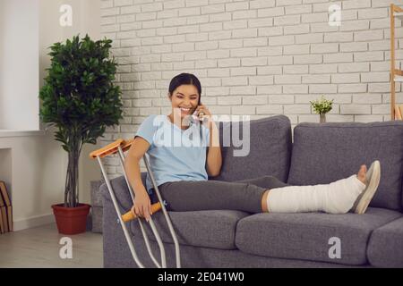 Giovane donna sorridente con gamba rotta seduta sul divano a. casa e parlare al telefono Foto Stock
