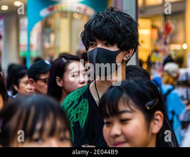 Un giovane mascherato con capelli neri scompigliati si trova schiacciato tra la folla su Takeshita-dori. La sua maglia scura è stampata con un motivo di drago verde. Harajuku, Tokyo, Giappone. Foto Stock