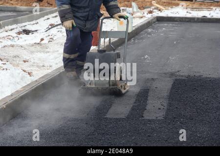 Il lavoratore usa il compattatore a piastre vibranti per rampare l'asfalto. Uomini al lavoro. Posa su strada di una nuova pavimentazione asfaltata. Foto Stock