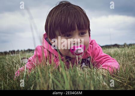 bambina sorridente con succhietto, espressione felice. Adagiato sull'erba in un parco. Mostra di sentimenti felici. Foto Stock