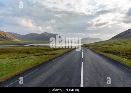 Deserta strada di montagna che scende verso l'oceano in Islanda in una serata estiva nuvolosa Foto Stock