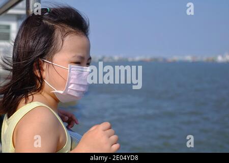 Una ragazza asiatica carina con una maschera chirurgica per proteggerla dall'epidemia di Covid-19, in attesa di una barca zattera vicino al porto, cercando di attraversare il fiume con lei Foto Stock