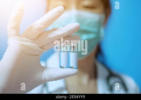 Una donna asiatica con maschera chirurgica e guanti di gomma bianca in una clinica, che contiene due bottiglie di vetro identiche di vaccino con etichetta bianca vuota Foto Stock