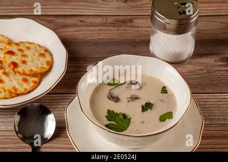 Vista laterale della zuppa cremosa di funghi decorata con foglie di prezzemolo è in una tazza artigianale di porcellana su un piatto piatto su un tavolo di legno servito con cracker, Foto Stock