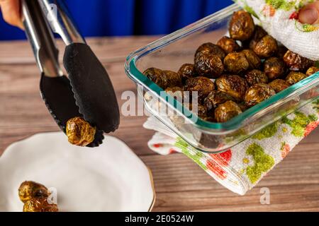 Primo piano immagine isolata di una spessa coppa di vetro di I germogli di Brussel cotti e stagionati vengono tenuti con un panno da cucina e servito in piatto di porcellana su Foto Stock