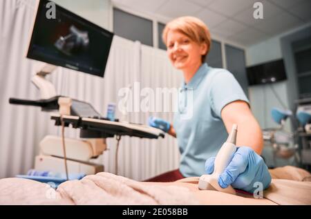 Concentrarsi sulla mano dell'ecografo femminile in guanti sterili che esaminano il paziente con uno scanner a ultrasuoni. Medico che esegue la scansione a ultrasuoni in cabina ginecologica. Concetto di diagnostica a ultrasuoni. Foto Stock