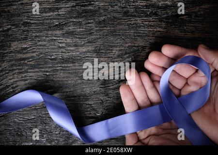 Vista dall'alto della mano maschio che tiene il nastro blu perivinkle su sfondo di legno con spazio di copia. Consapevolezza del cancro dello stomaco e dell'esofago, disturbi alimentari Foto Stock