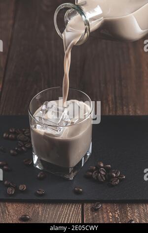 Versando il liquore di caffè Cream in un bicchiere di ghiaccio, circondato da chicchi di caffè su sfondo di legno scuro. Foto Stock
