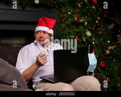 Un malato da solo saluta il nuovo anno. Un uomo con un computer portatile accanto a un albero di Natale. Foto Stock