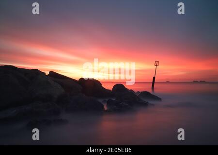 Una lunga esposizione cattura striature di colore fuoco nel cielo nuvoloso mentre il sole sorge sul mare al largo della spiaggia di Bournemouth. Foto Stock