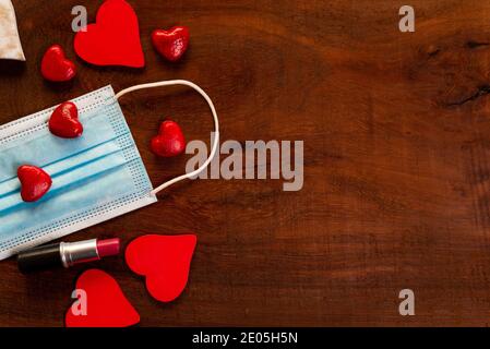 concetto di igiene del giorno di covid valentine con spazio copia. maschera facciale con cuori rossi su sfondo ligneo. posa piatta con rossetto per la celebrazione dell'amore Foto Stock