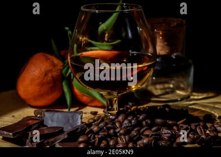 Bicchiere di brandy su tavolo di legno con cioccolato, caffè e mandarini stretti. Foto Stock