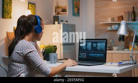 Creatore di contenuti freelance che lavora straordinario da casa per rispettare la scadenza. Donna videografa montaggio audio film montaggio su portatile professionale seduta su scrivania in cucina moderna a mezzanotte Foto Stock