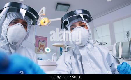 Squadra dentale che si appoggia sopra il paziente per lavorare sulle gengive del paziente vestite in abito di nocciolo in studio dentale con nuovo normale. Team di stomatologia che indossa attrezzature di sicurezza contro il coronavirus trattamento dei denti problema. Foto Stock