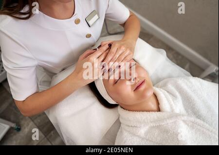 Una bella giovane donna che si rilassa su un tavolo da massaggio mentre Specialista della bellezza che effettua un massaggio lifing del viso nel salone SPA Foto Stock