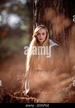 Naturalmente bella giovane donna che guarda direttamente alla macchina fotografica in un'immagine editoriale di moda ambientato in una foresta che indossa un cappotto bagnata dal sole d'inverno glorioso. Foto Stock