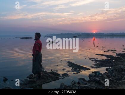 Pescatore in piedi su rocce sulla riva all'alba al lago Taungthaman, Amarapura, Mandalay, Myanmar (Birmania), Asia nel mese di febbraio Foto Stock