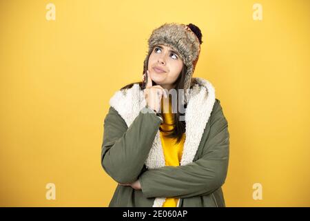 Giovane bella donna che indossa un cappello e un inverno verde cappotto su sfondo giallo pensando e guardando al lato Foto Stock