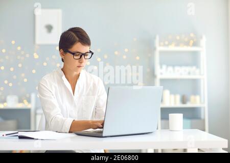 Giovane donna di affari in occhiali che pungono e che lavora su un computer portatile online da casa Foto Stock