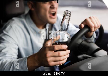 vista tagliata di un uomo aggressivo e ubriaco con una bottiglia di whisky che grida durante la guida dell'auto Foto Stock