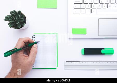 Posto di lavoro in ufficio con scrittura a mano in notebook, laptop e materiali da ufficio ecologici su una scrivania bianca. Foto Stock