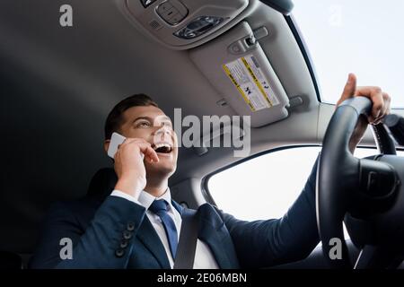 Vista ad angolo basso di un uomo positivo in tuta che parla telefono cellulare durante la guida dell'auto Foto Stock