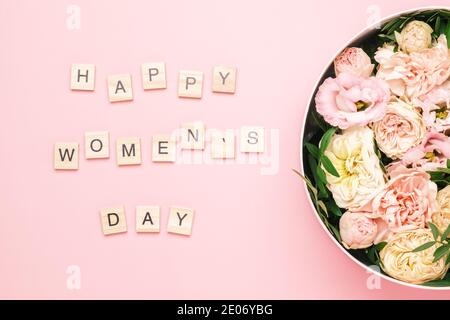 Happy Womens day lettering sulle piazze di legno con lettere sullo sfondo rosa e grande scatola rotonda con fiori rossi e rose. Foto Stock