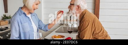 vista laterale della moglie felice anziana che alimenta il marito con pomodoro in ciliegio in cucina, striscione Foto Stock