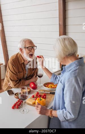 vista posteriore della moglie anziana che alimenta felice marito con ciliegia pomodoro vicino al tavolo con cibo in cucina Foto Stock