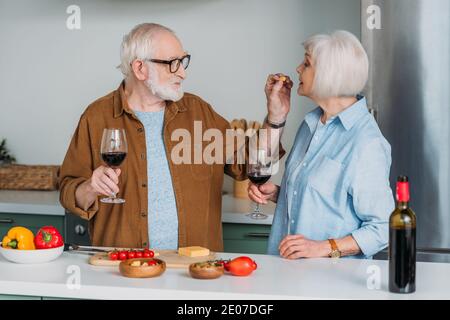 uomo anziano sorridente con bicchiere di vino che alimenta la moglie con il pezzo di formaggio in cucina su sfondo sfocato Foto Stock